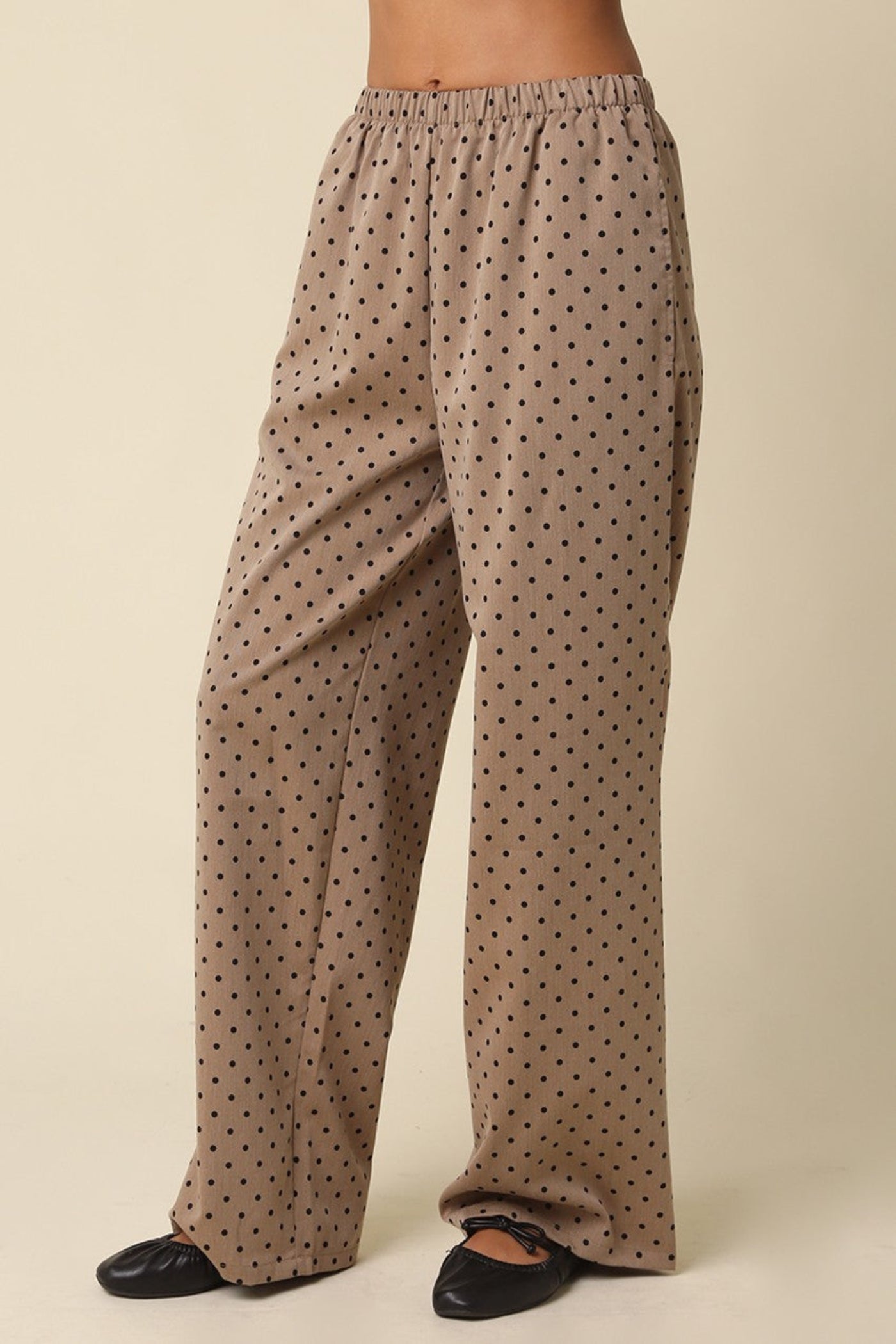 A person wearing loose, wide-leg pants in a warm taupe-beige color with a small black polka dot pattern, paired with black flats, shown against a plain beige studio background.