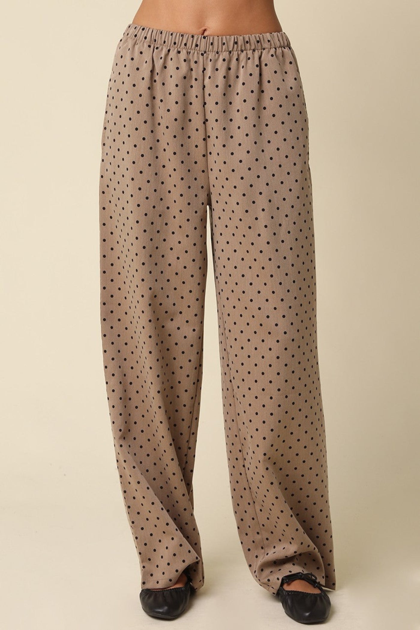 A person wearing loose, wide-leg pants in a warm taupe-beige fabric with a small black polka-dot pattern, paired with black flats, shown against a plain beige studio background.