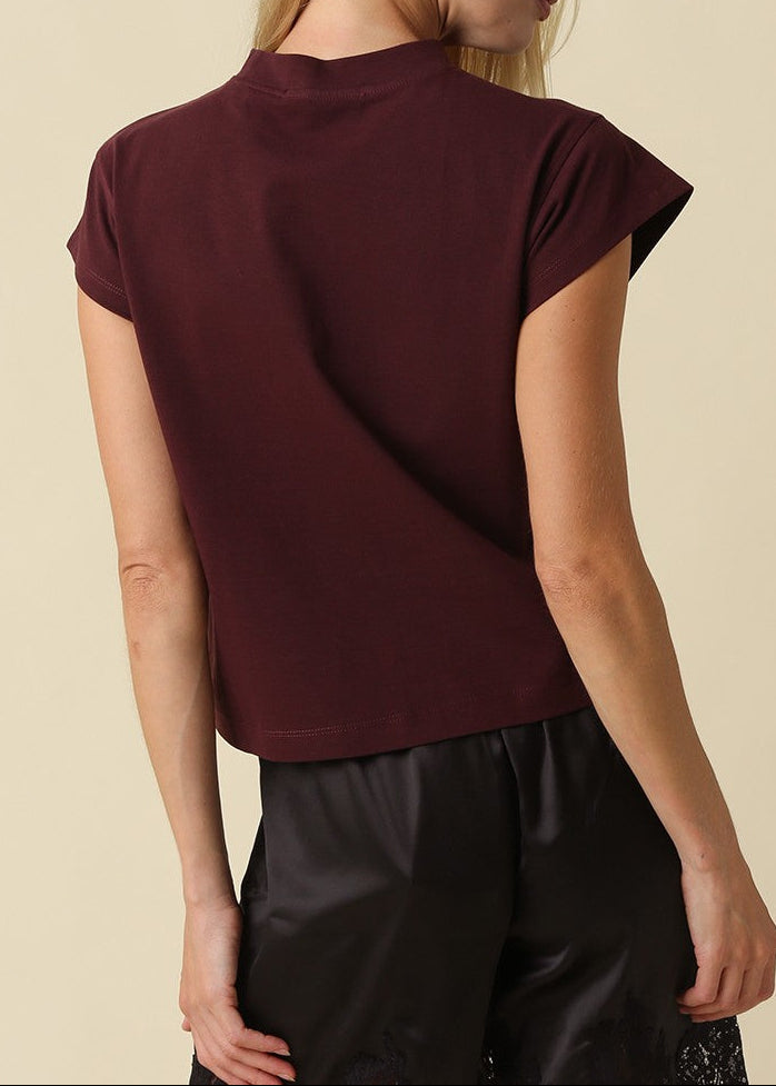 A woman shown from the back wearing a short-sleeve crewneck t-shirt in deep burgundy, paired with black pants, against a solid beige background.