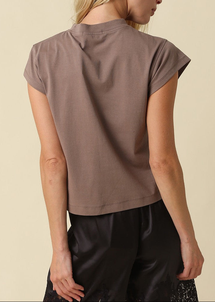 A woman shown from the back wearing a taupe short-sleeve crop top and black pants, photographed against a solid light beige background.