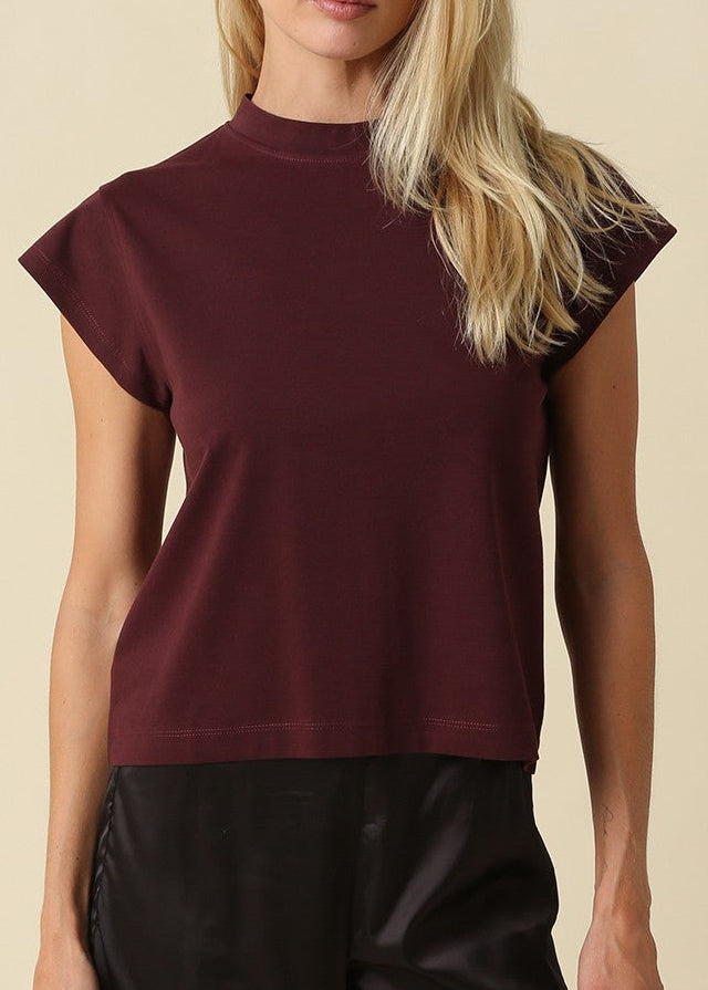 A blonde woman modeling a short-sleeve mock neck top in burgundy, paired with black pants, against a solid beige background.