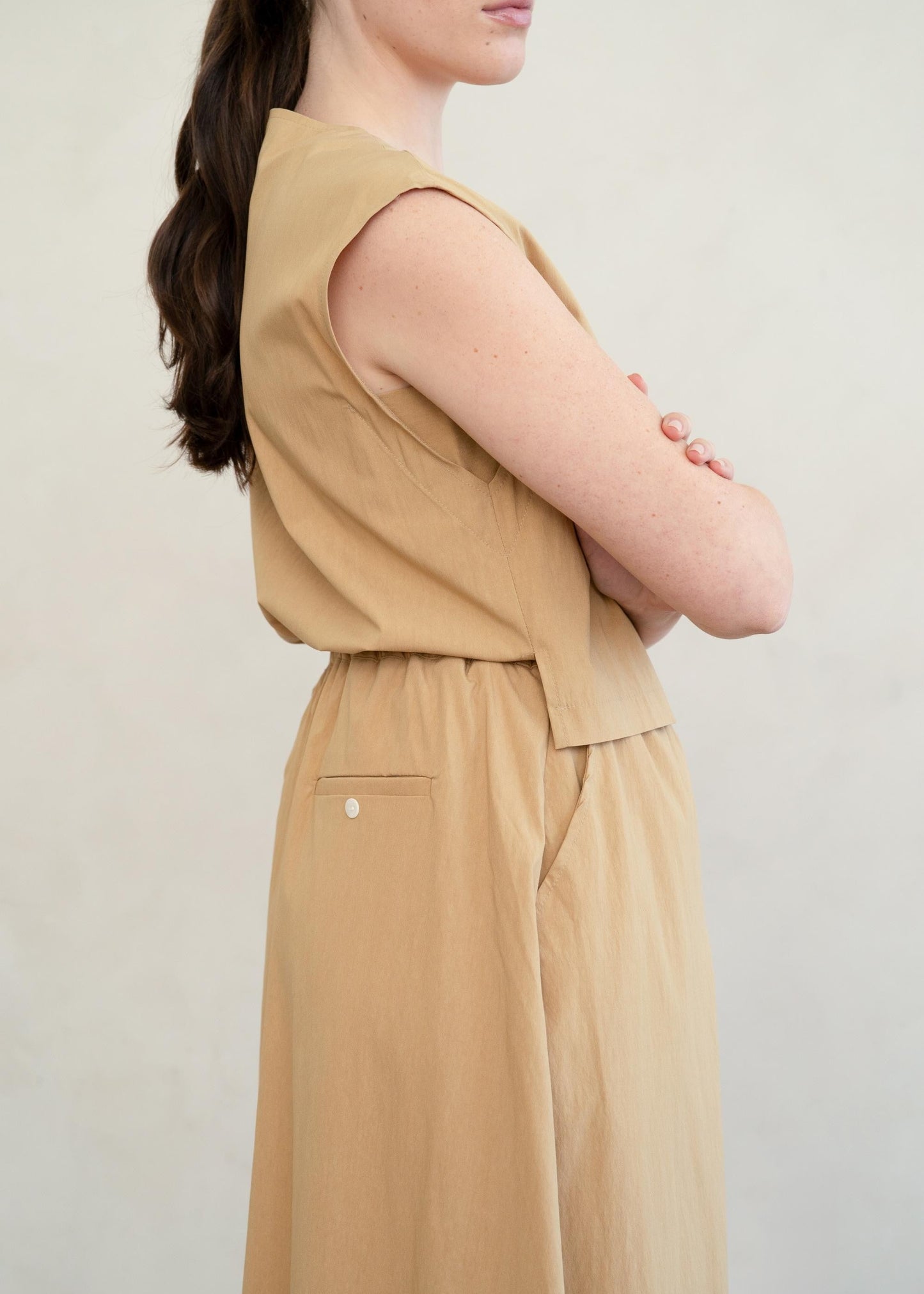 A woman wearing a sleeveless tan jumpsuit, shown from the side against a plain light background.