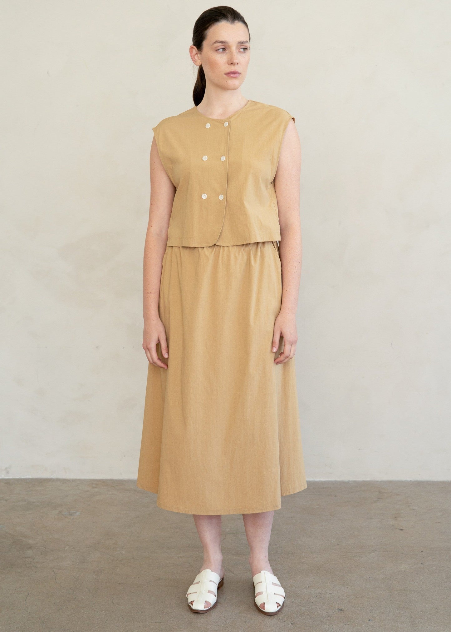 A woman modeling a sleeveless tan midi dress with a buttoned cropped bodice and a flared skirt, standing against a plain light beige wall.