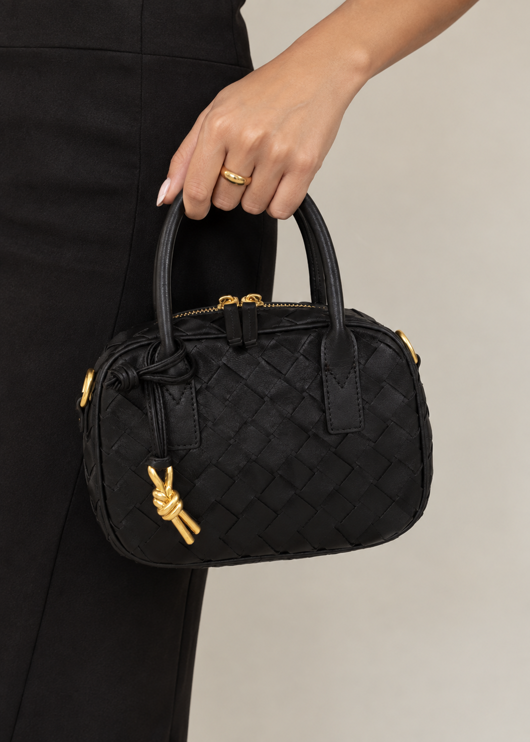 A close-up of a person holding a small black woven leather handbag with gold-tone hardware and a knotted charm, against a plain light gray background.