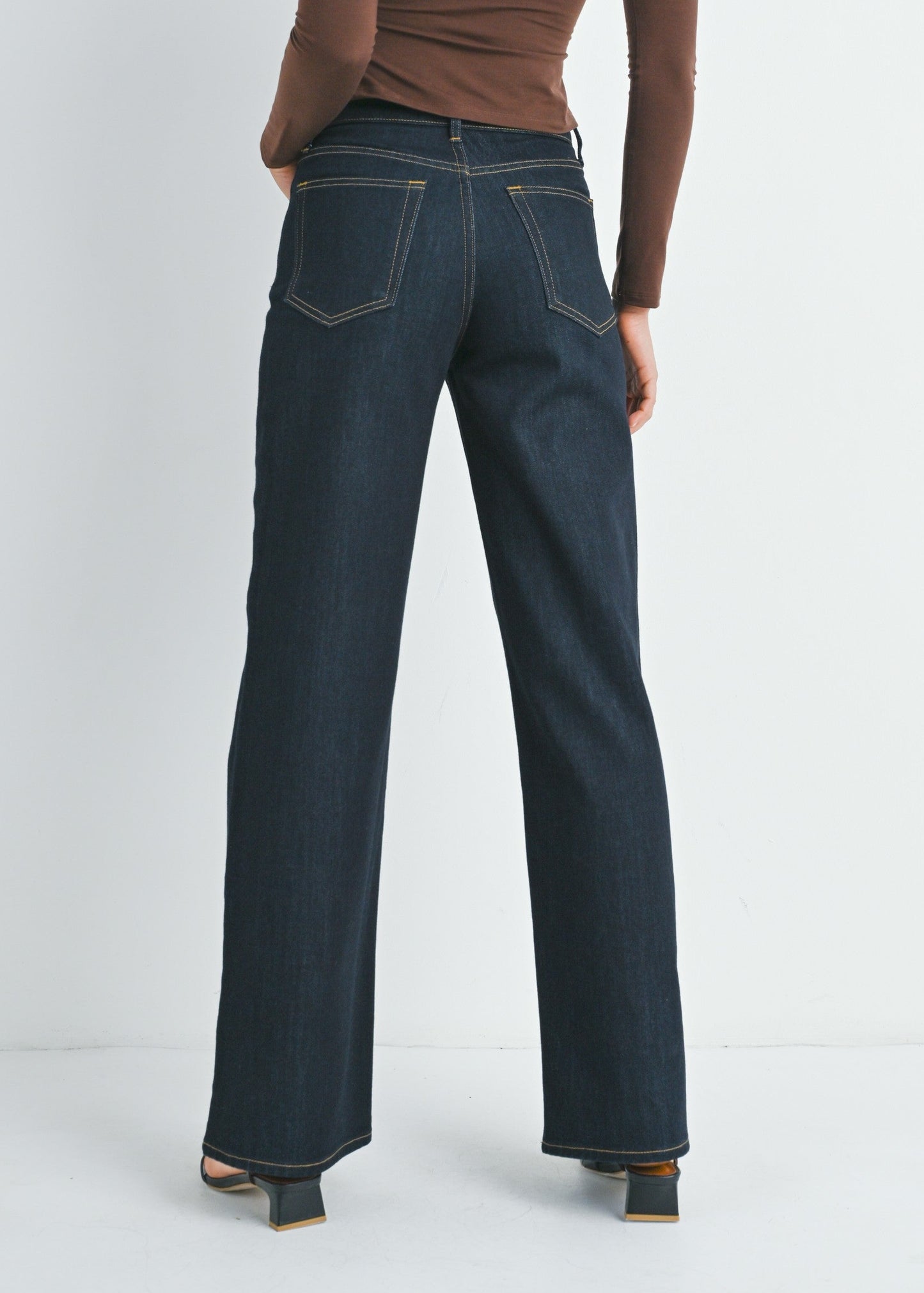 A woman is shown from the back wearing a brown off-the-shoulder long-sleeve top and dark blue wide-leg jeans against a plain light gray studio background.