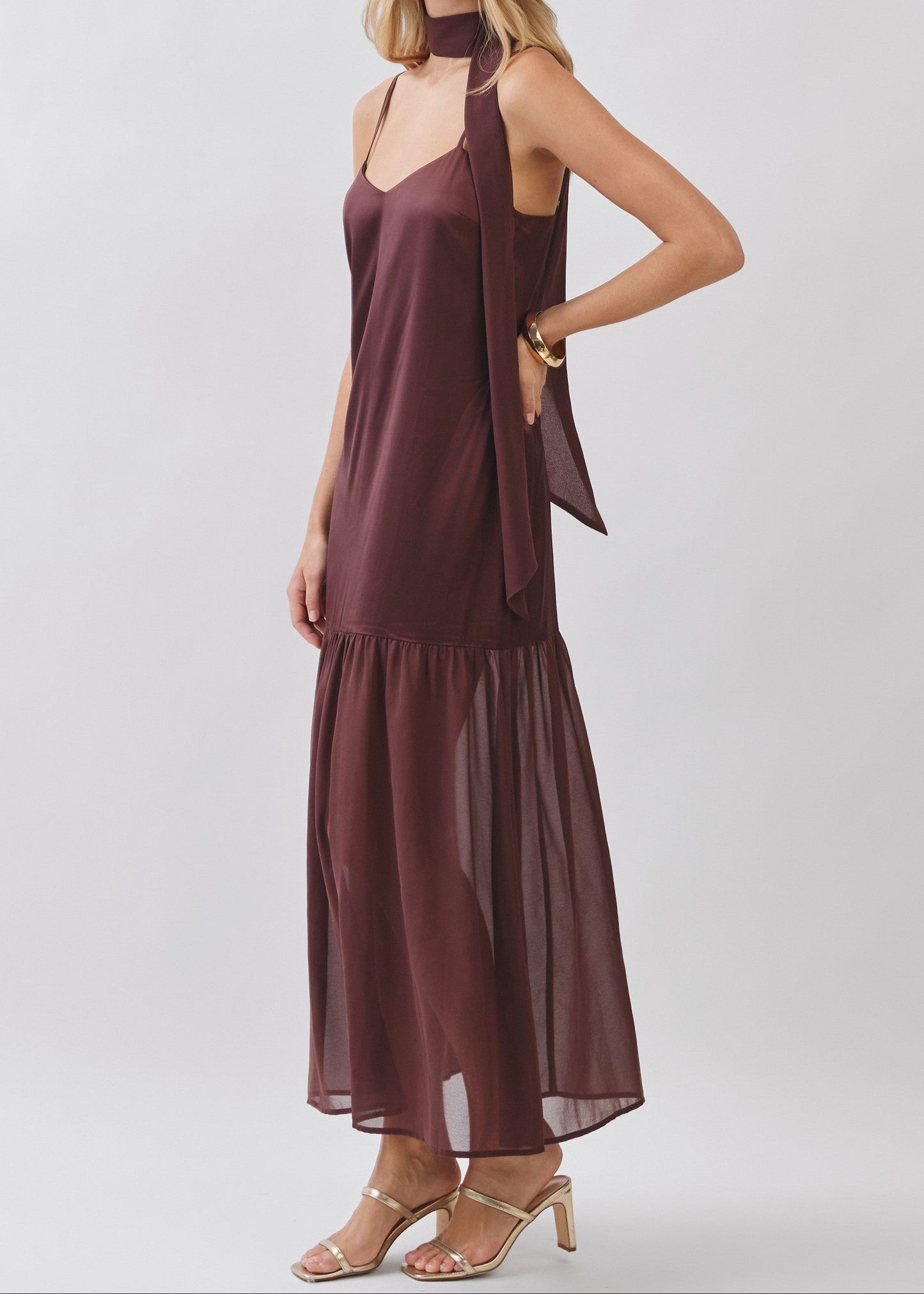 A woman modeling a sleeveless, high-neck maxi dress in a deep plum-brown color. The dress features a sheer, layered ruffled hem and a draped side detail.