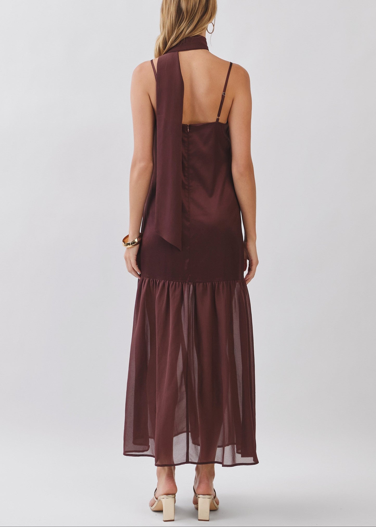 A woman viewed from the back wearing a sleeveless dark brown maxi dress with a draped halter-style back detail and thin straps, paired with beige block-heel sandals against a light gray studio background.