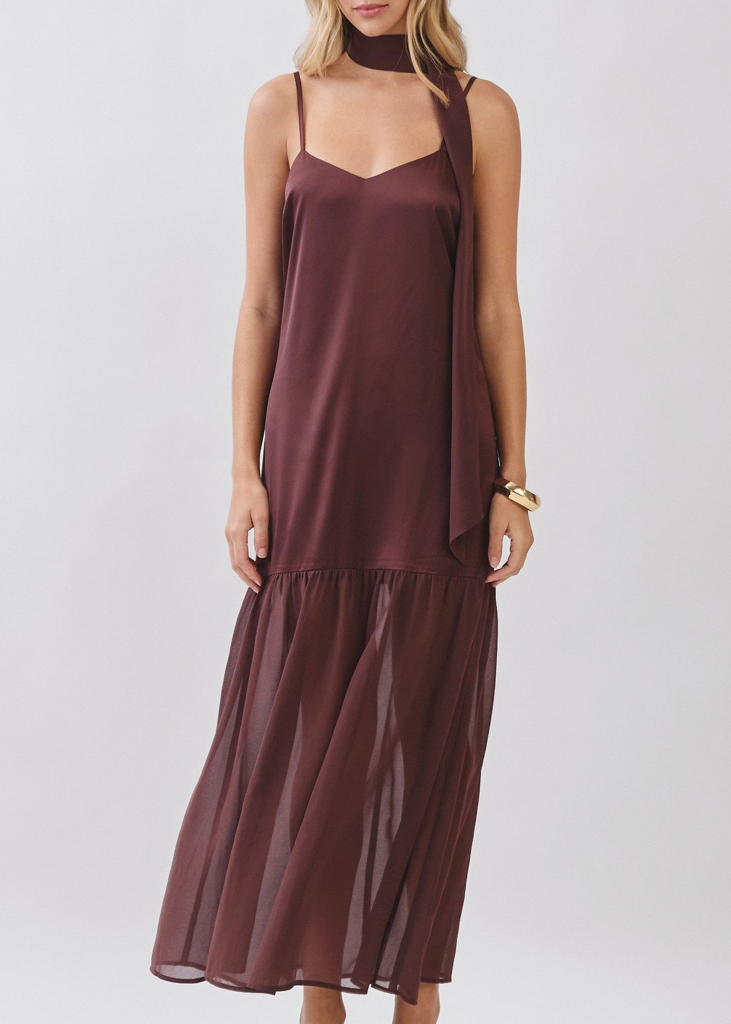 A woman modeling a sleeveless, high-neck, tiered maxi dress in a deep plum-brown color, paired with gold-toned sandals and a gold bracelet, against a light gray studio background.