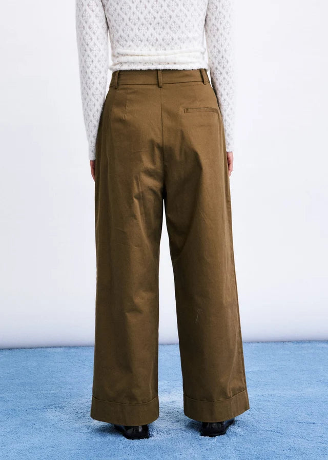 A person shown from the back wearing wide-leg brown trousers, a white textured long-sleeve top, and black shoes, standing on a blue surface against a white wall.