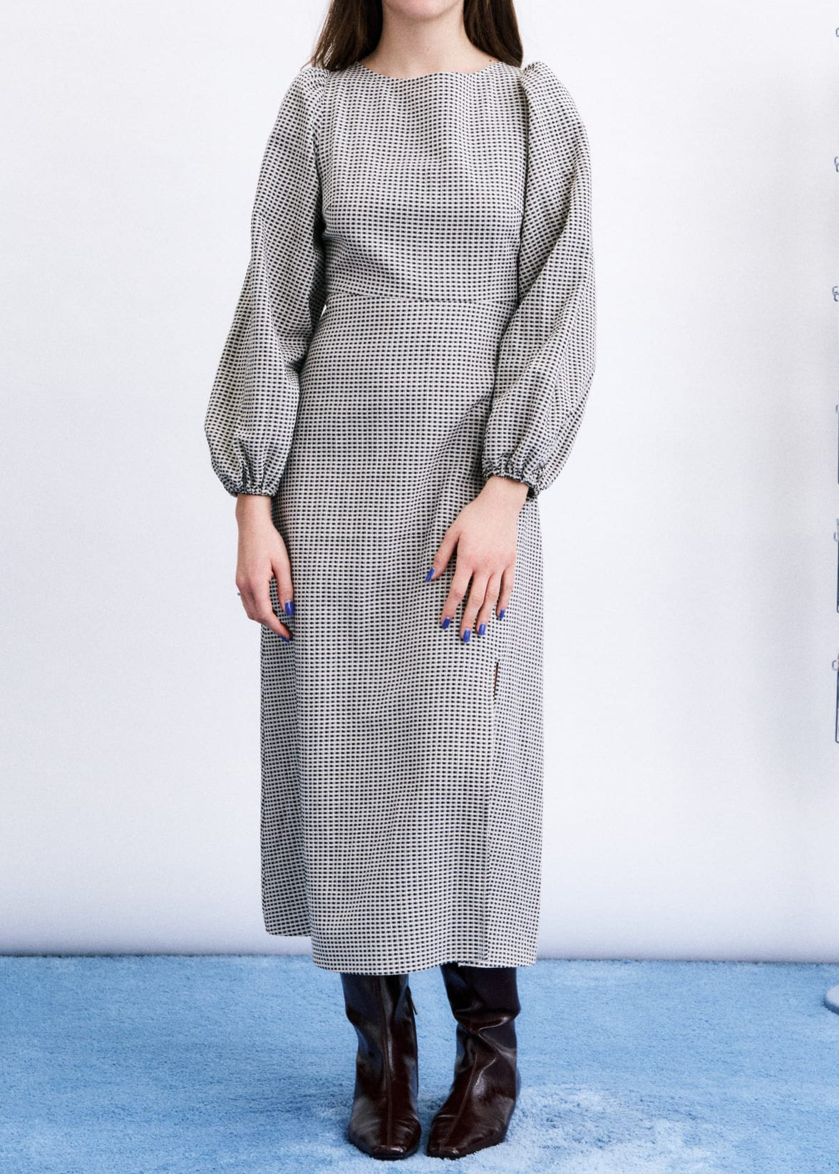 A woman stands in a studio wearing a long-sleeved gray and white micro-check dress with puff sleeves and dark brown ankle boots. A light blue floor and a white wall are visible, with a magazine rack on the right.