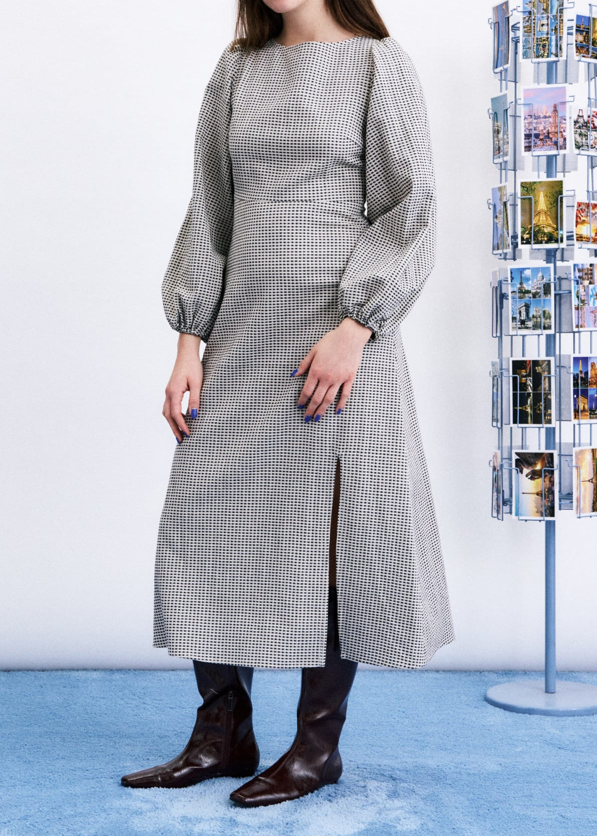 A woman models a long-sleeved, knee-length gray and white checkered midi dress with a front slit, paired with dark brown boots, standing in front of a white wall and a blue carpet. A modern rotating photo display stand is visible on the right.