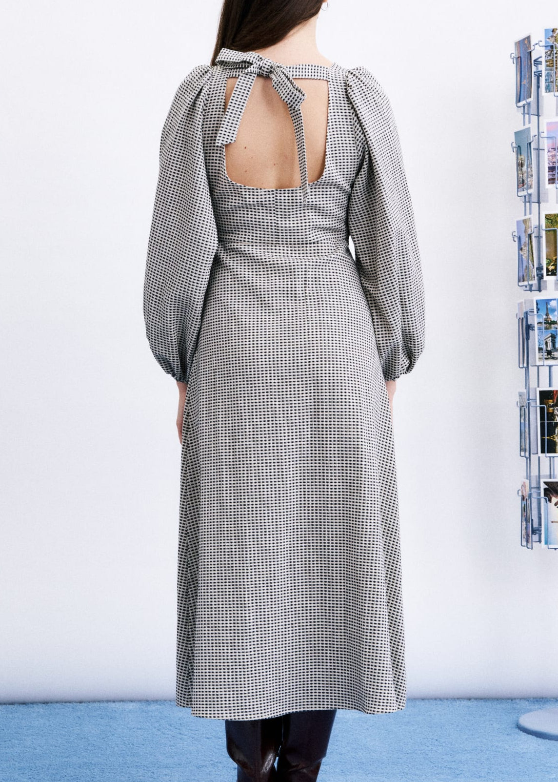 A woman stands with her back to the camera wearing a long-sleeved gray and white gingham midi dress with a tie-back neckline and puff sleeves. She is standing on a light blue carpet next to a white wall with a magazine rack.