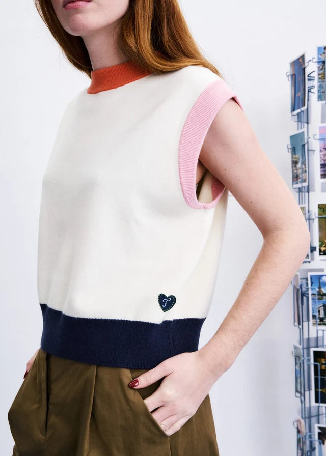 A woman wearing a color-block knit sweater vest with an off-white body, a navy blue hem, a pink sleeve, and a rust orange mock neck. She is wearing olive brown trousers, standing next to a wall display.