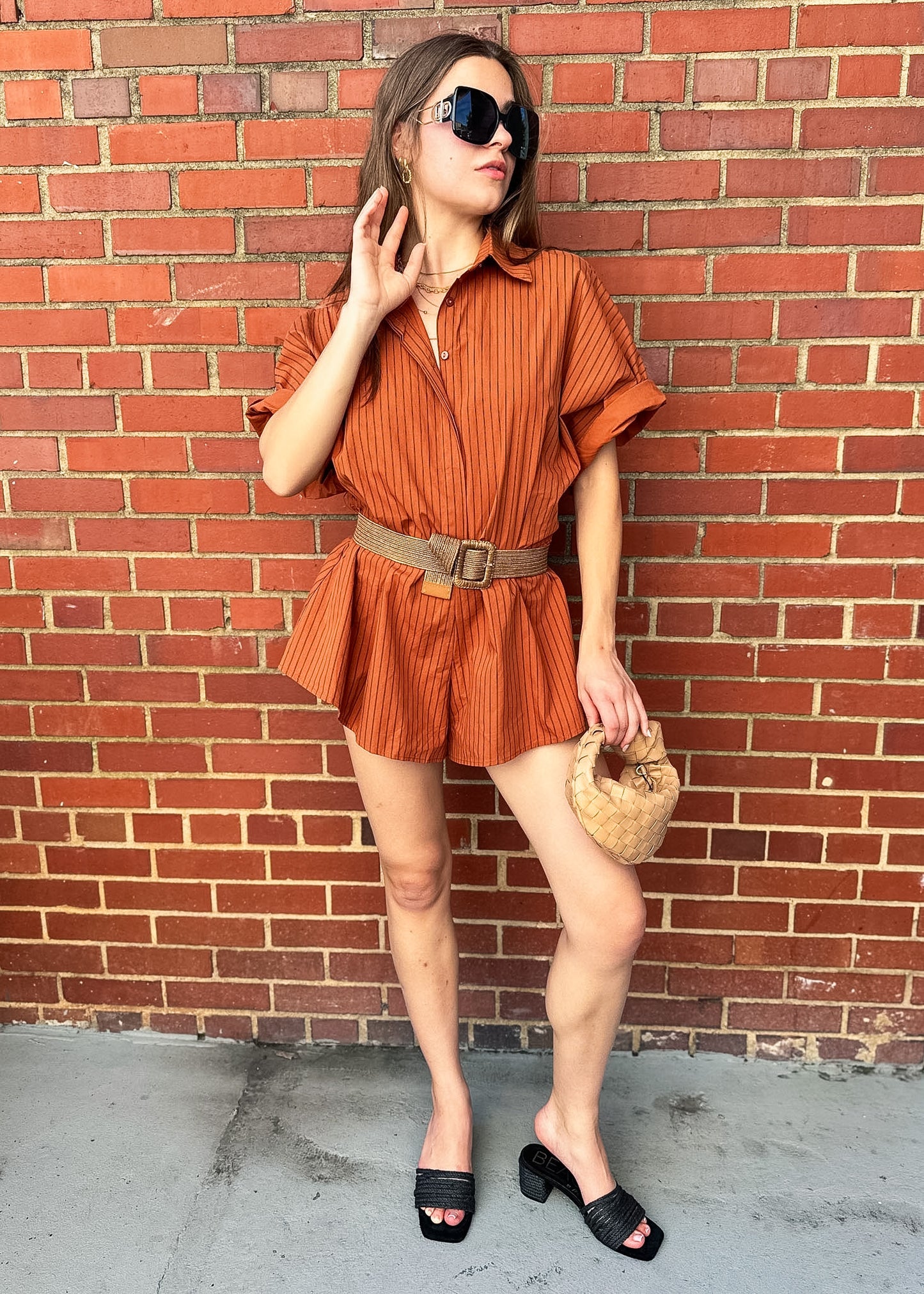 A woman stands against a red brick wall wearing a rust-orange vertically striped short-sleeve romper with a tan belt, black sunglasses, black slide sandals, and holding a woven straw handbag.
