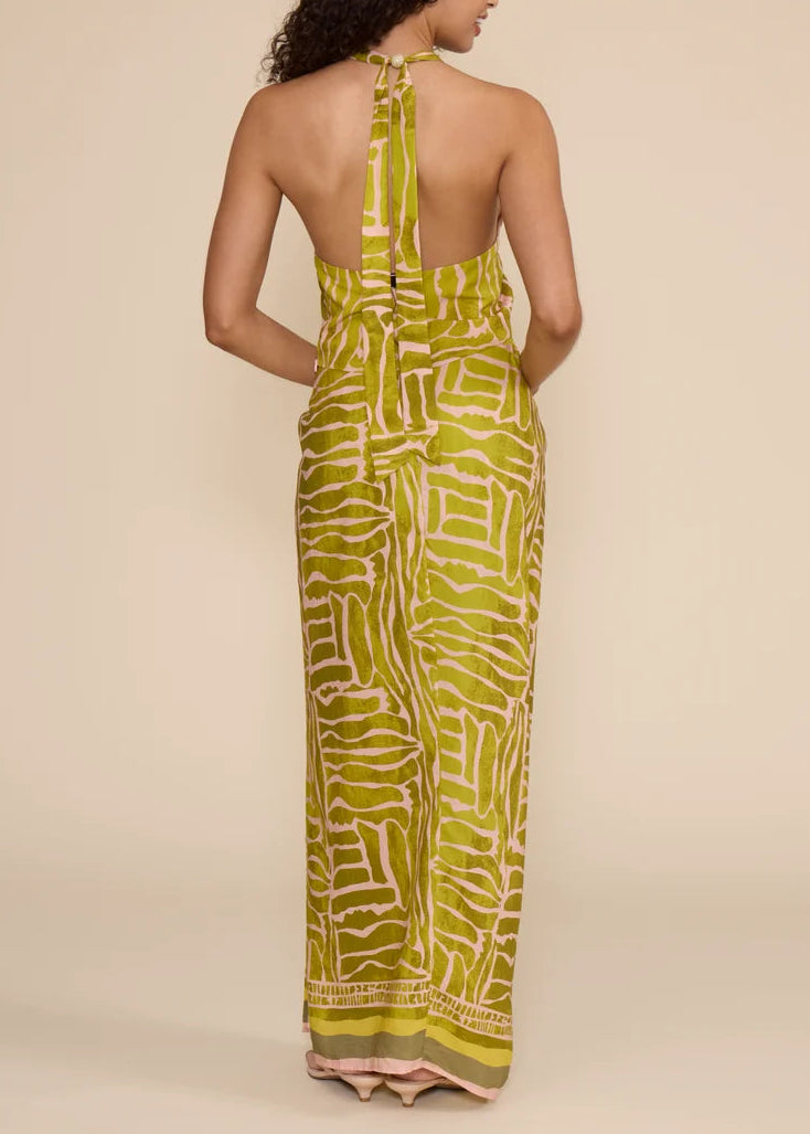 A woman shown from the back wearing a sleeveless halter-neck maxi dress with an open back and a bold abstract print in olive green and cream, set against a plain beige studio background.