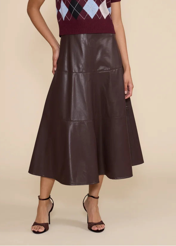 A model stands against a solid beige studio background wearing a short-sleeve knit top with an argyle pattern in burgundy, navy, light blue, and white, paired with a dark brown faux-leather midi skirt and black strappy heels.