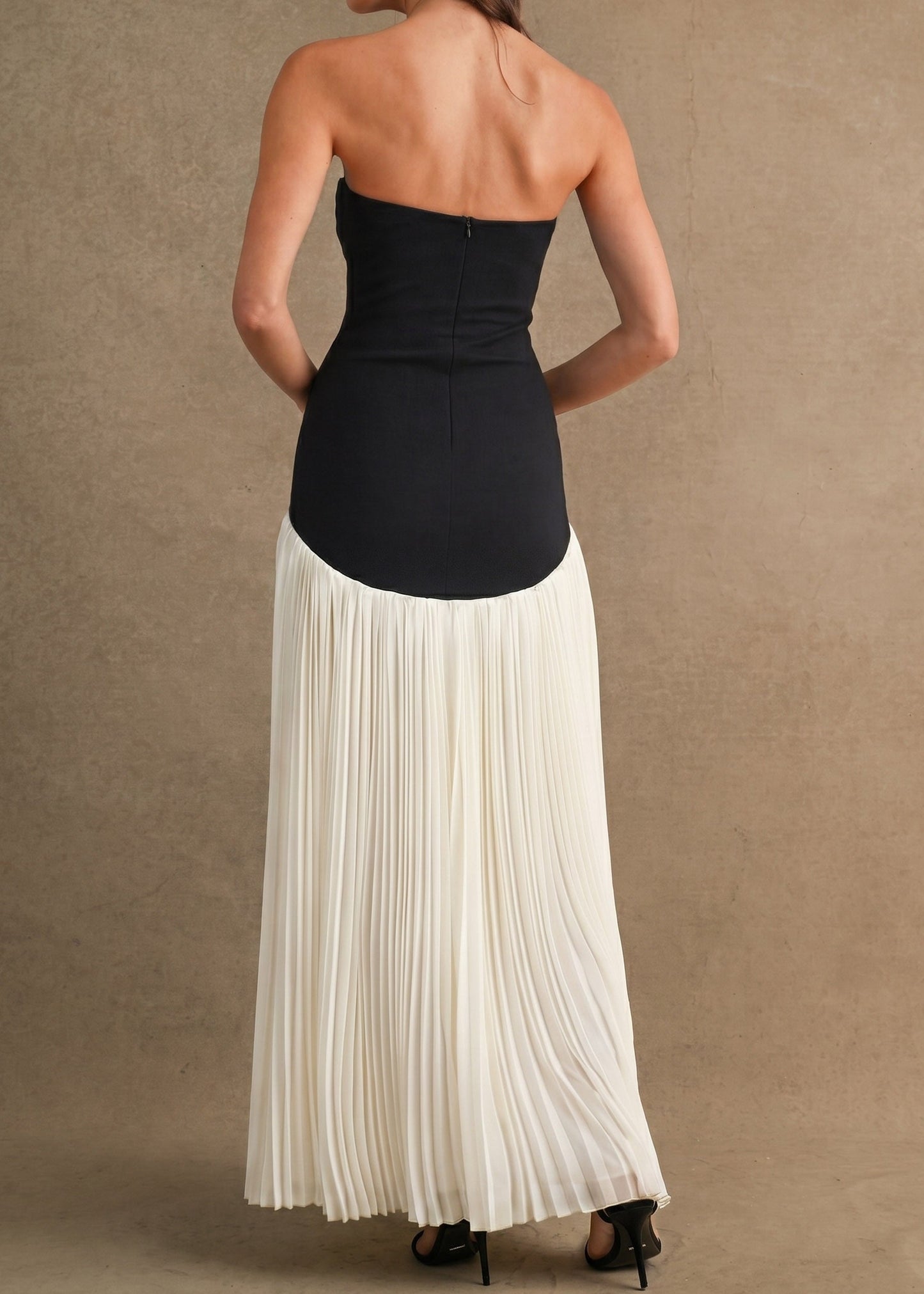 A woman shown from the back wearing a strapless maxi dress with a black fitted bodice and a long pleated off-white skirt, standing against a textured beige studio backdrop.