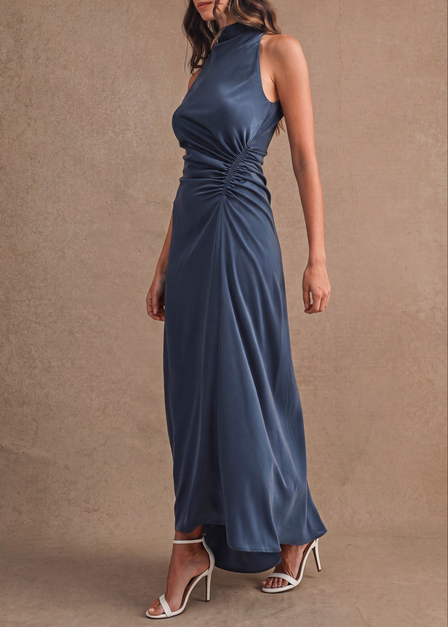 A woman modeling a sleeveless navy blue satin maxi dress with a gathered ruched waist, standing against a textured beige studio backdrop.