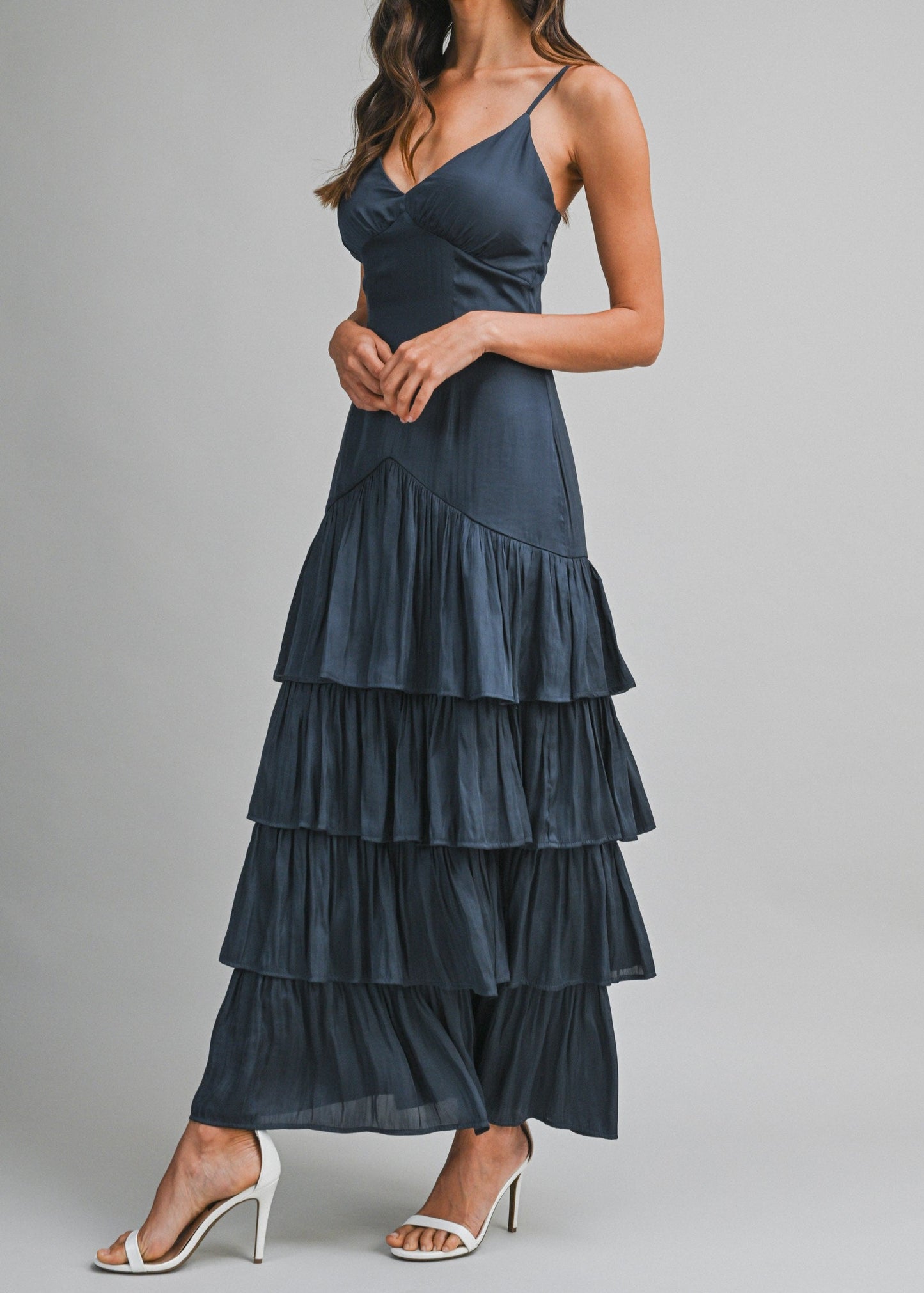 A woman modeling a dark slate blue tiered maxi dress with spaghetti straps and white high-heeled sandals against a solid gray studio background.