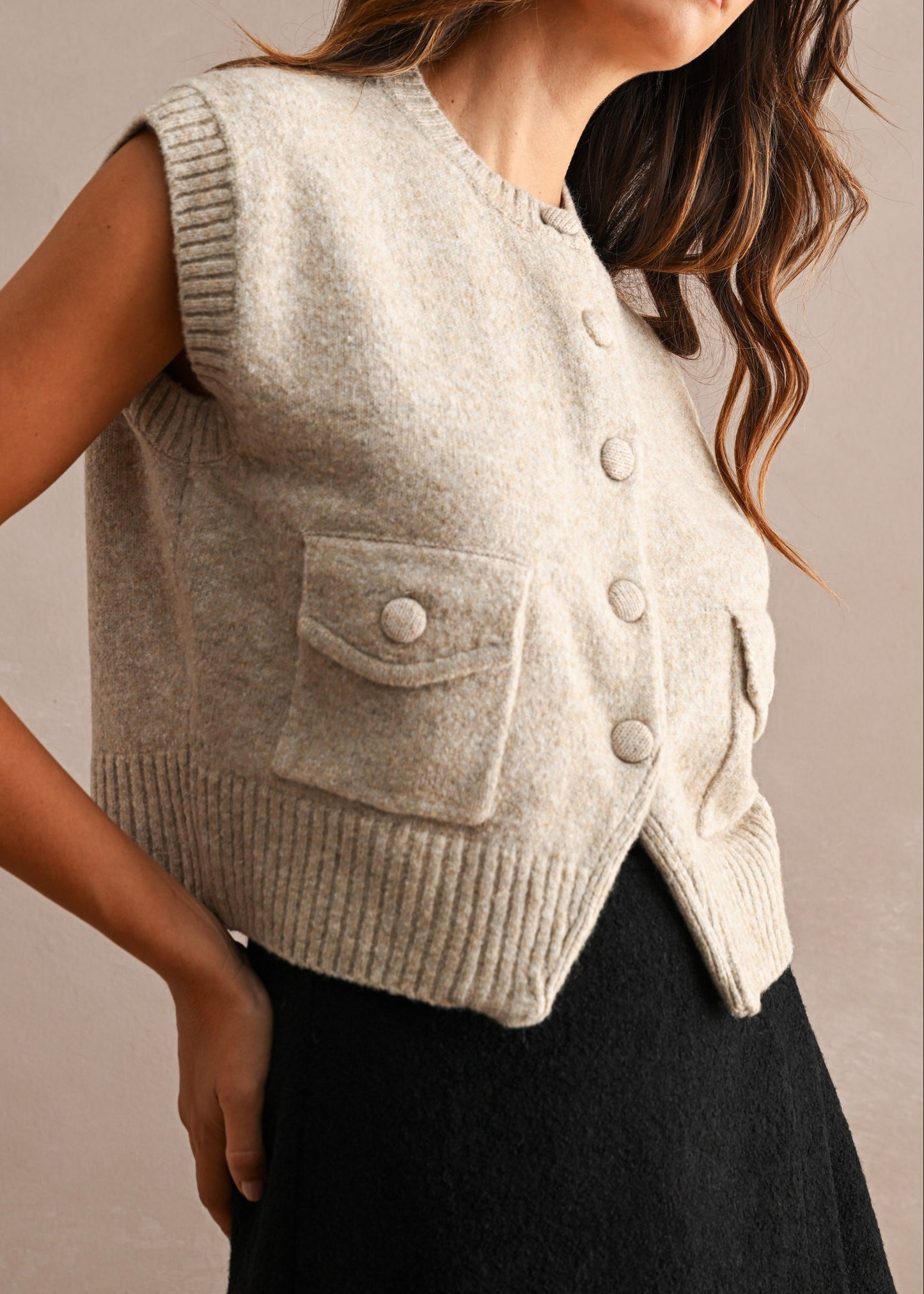 A woman wearing a light beige knit sleeveless vest with front buttons and flap pockets, paired with a black skirt, against a neutral beige background.