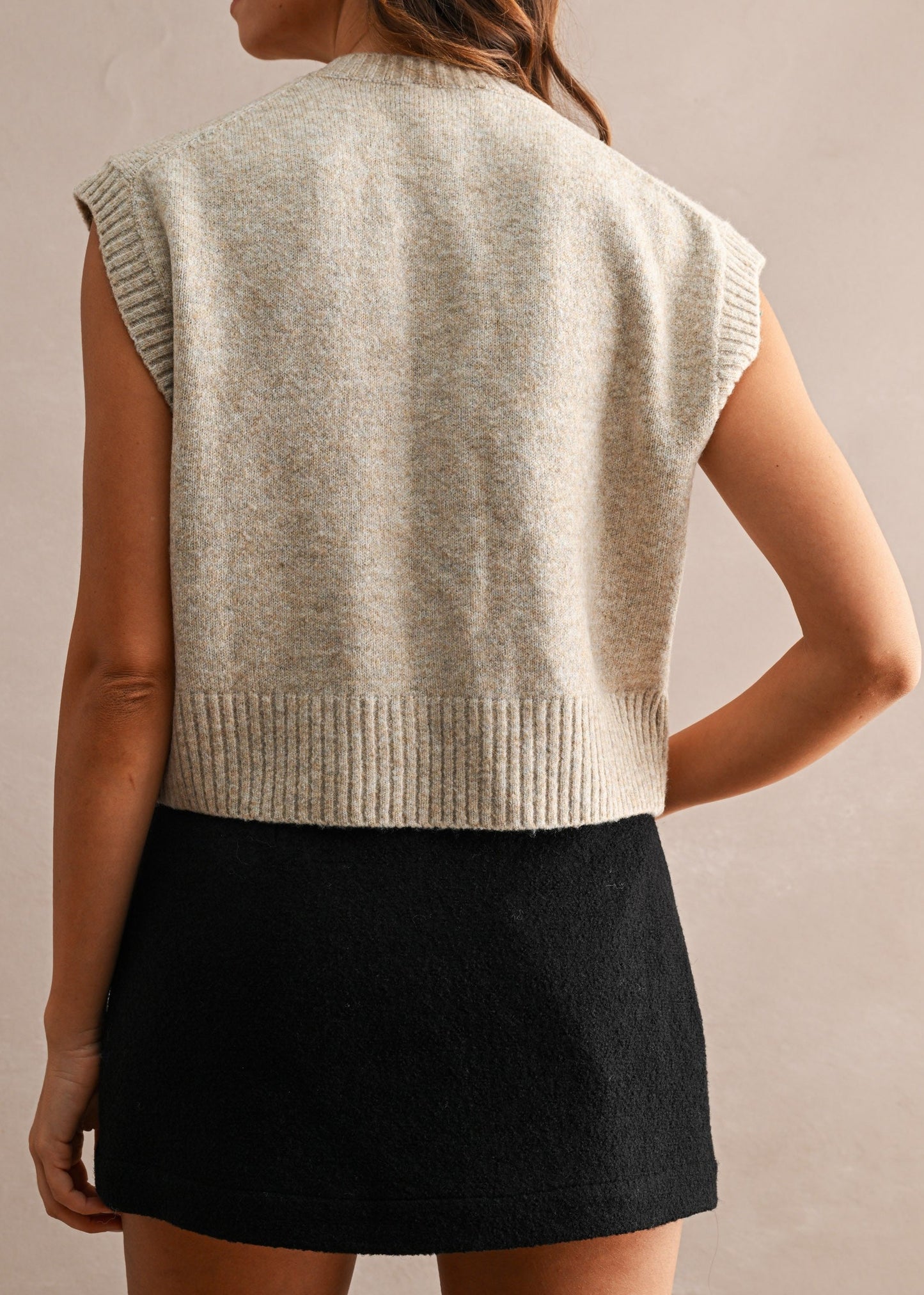 A woman shown from the back wearing a sleeveless knit sweater vest in a light oatmeal beige color, paired with a black mini skirt, against a neutral textured beige background.