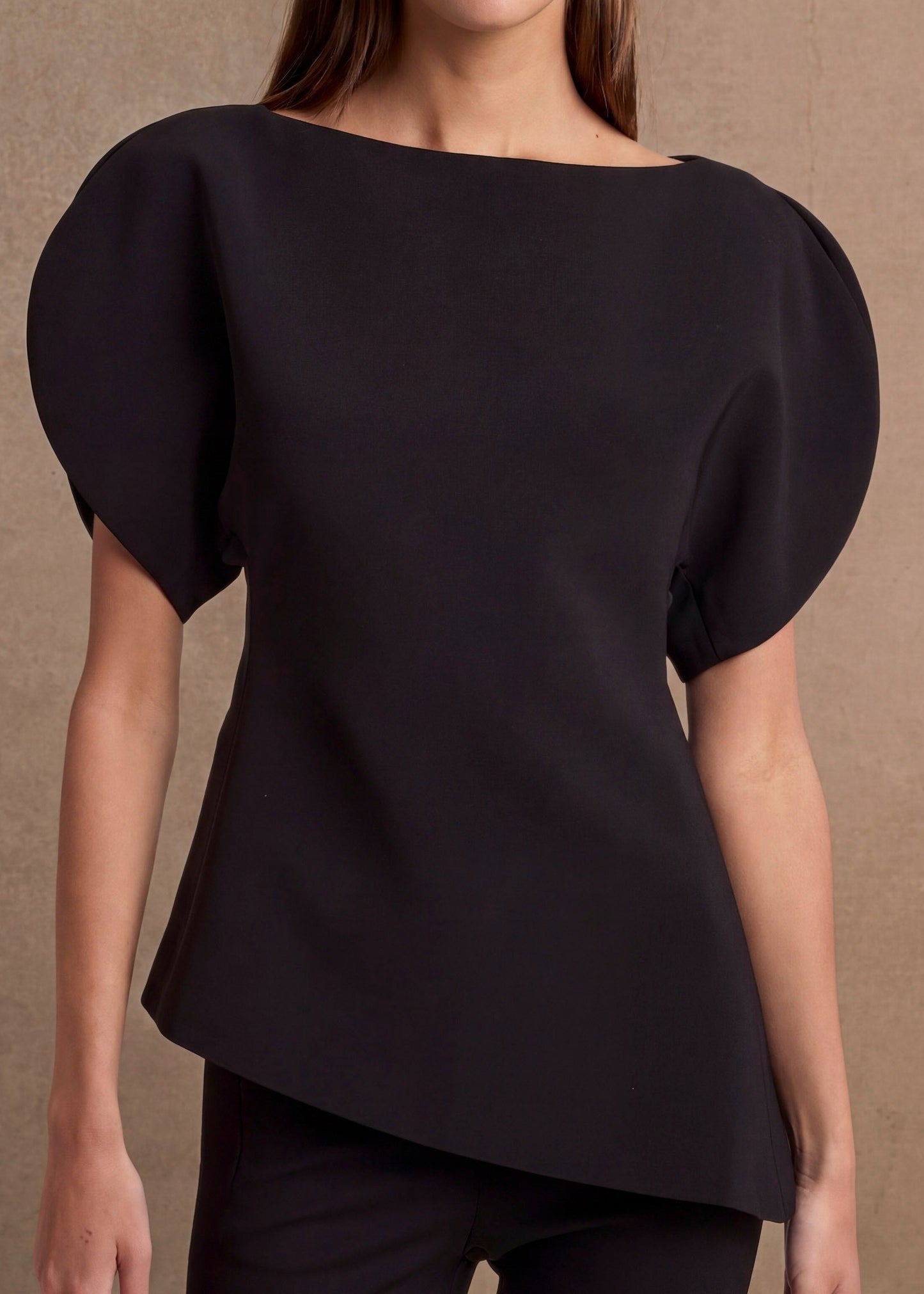 A woman modeling a black boat-neck top with voluminous puff sleeves, paired with black pants, set against a textured beige studio background.