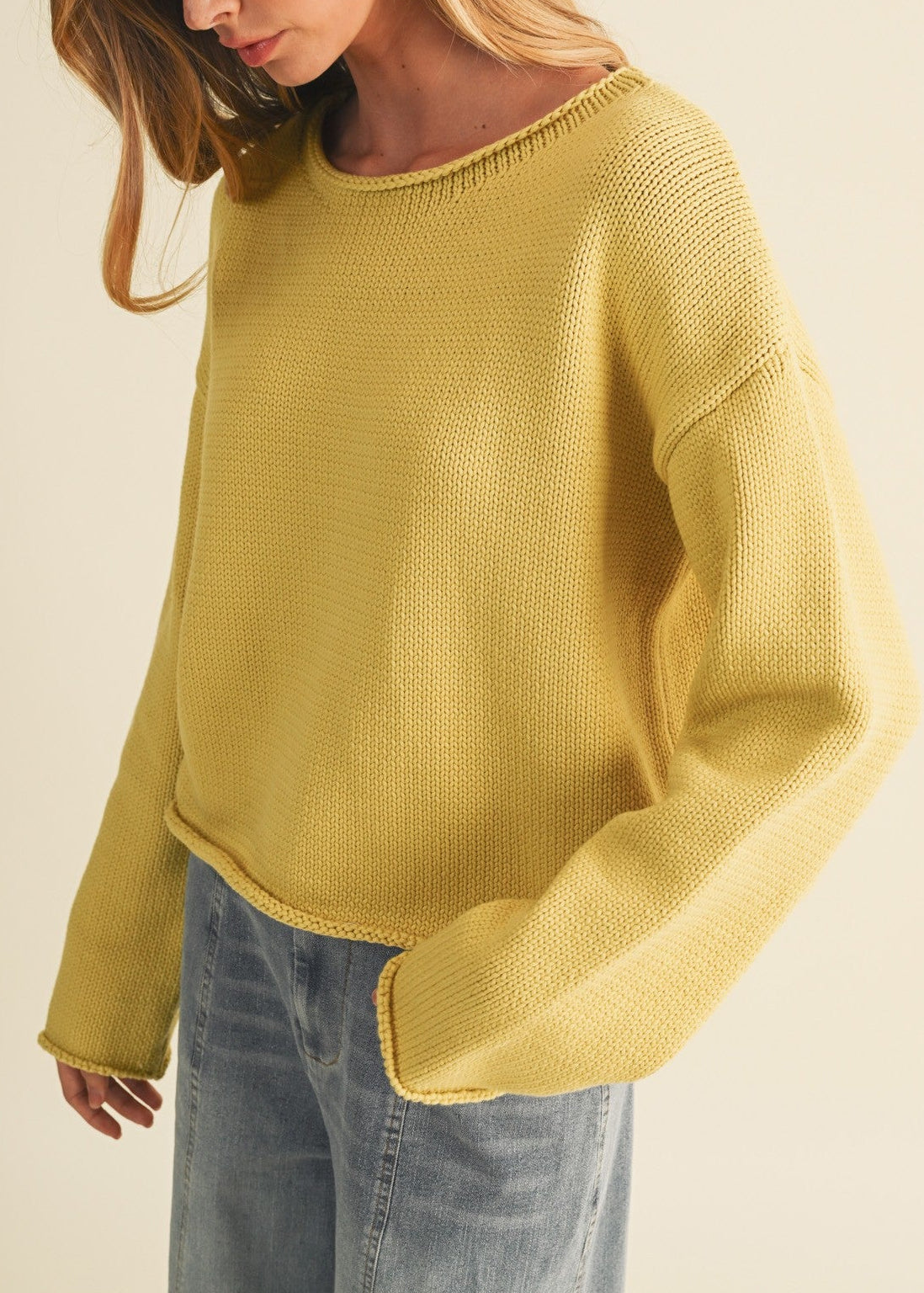 A woman wearing a textured mustard yellow knit sweater and light blue jeans, photographed against a plain beige studio background.