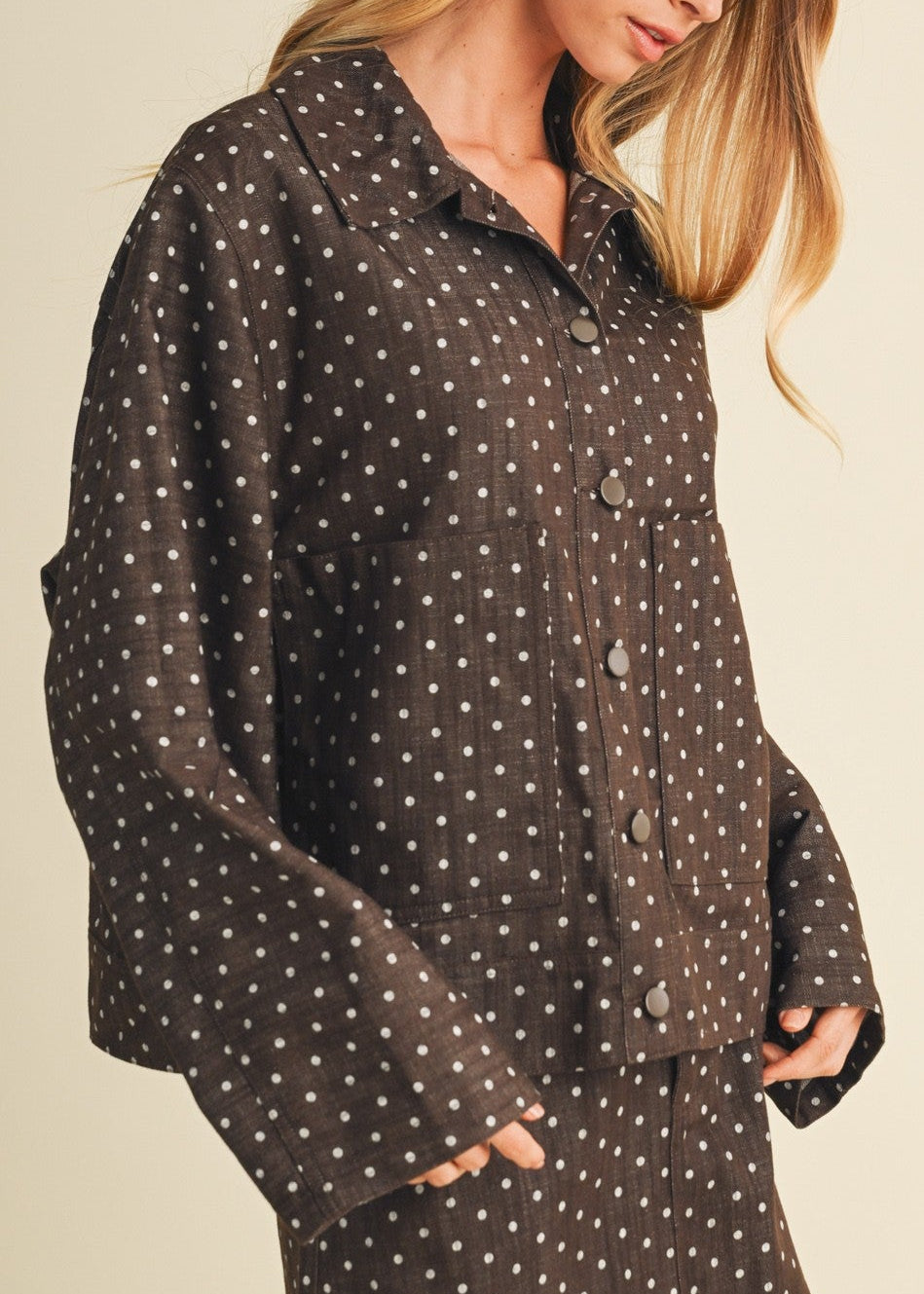 A woman wearing a dark brown button-up shirt and matching shorts set with a small white polka dot pattern, posed against a solid light beige background.