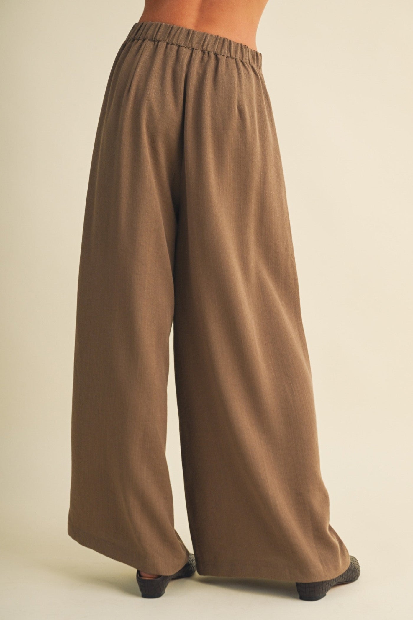 A person shown from the back wearing loose, wide-leg brown pants with an elastic waistband, photographed against a plain light beige studio background.