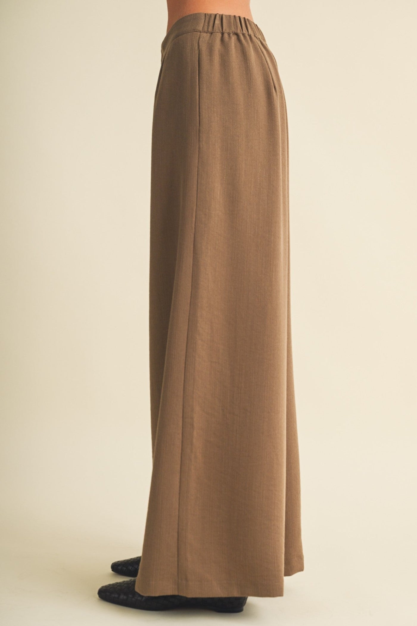 A person shown from the back wearing a long, wide-leg brown skirt or drape with an elastic waistband, photographed against a plain warm beige studio background.