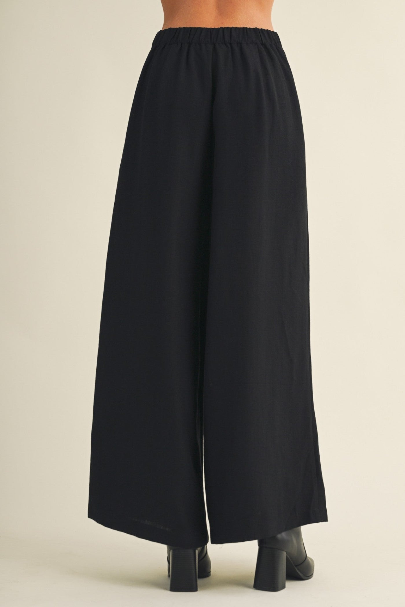 Black wide-leg cropped pants shown from the back on a model, featuring an elastic waistband and a straight, flowing silhouette, paired with black block-heeled boots against a light beige background.
