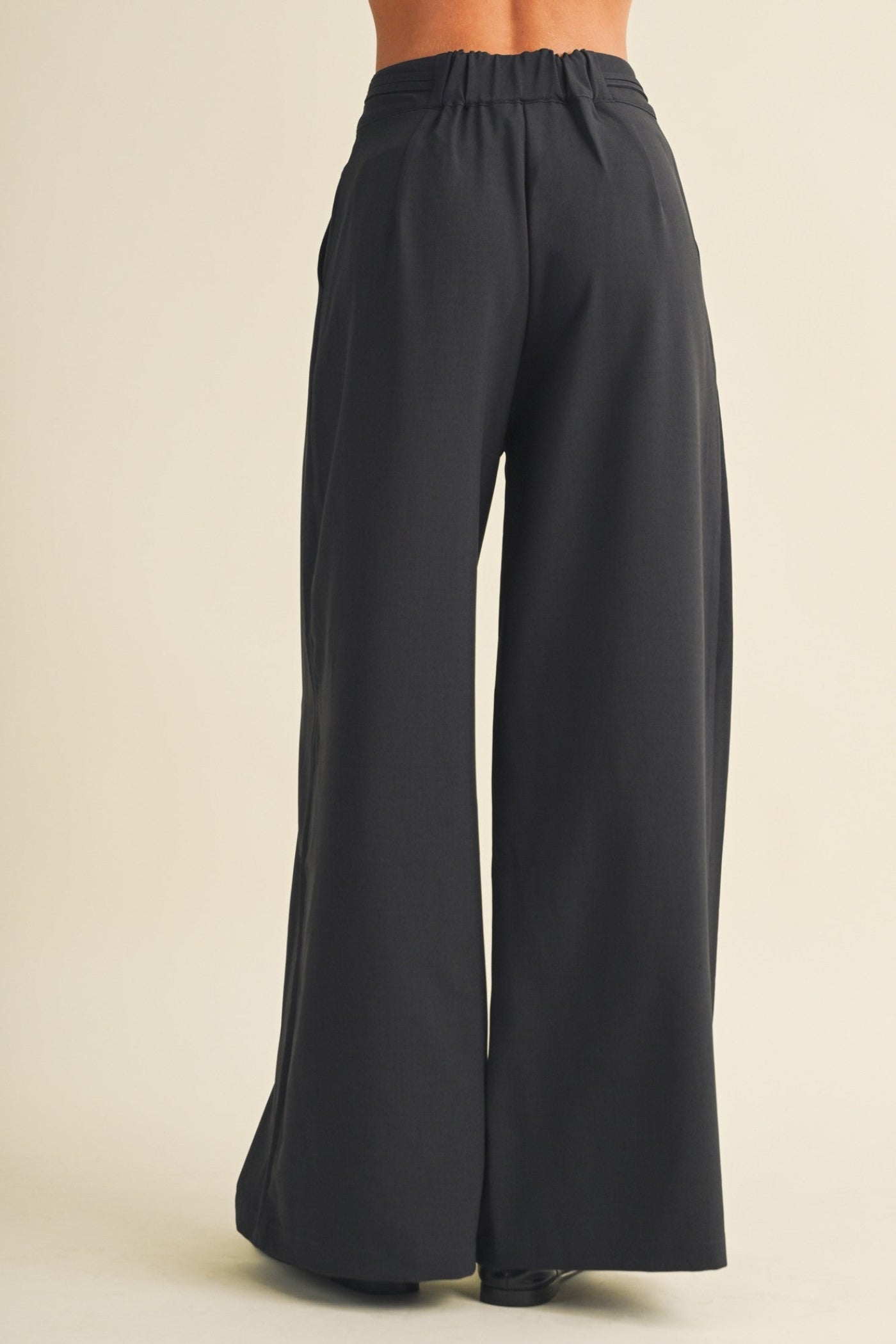 Back view of a model wearing black wide-leg trousers with an elastic waistband, set against a solid light beige background.