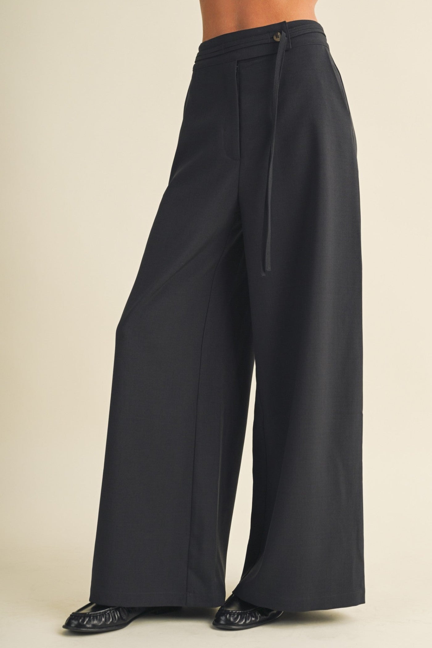 Black wide-leg trousers with a drawstring waistband, shown on a model from the waist down against a plain beige studio background.