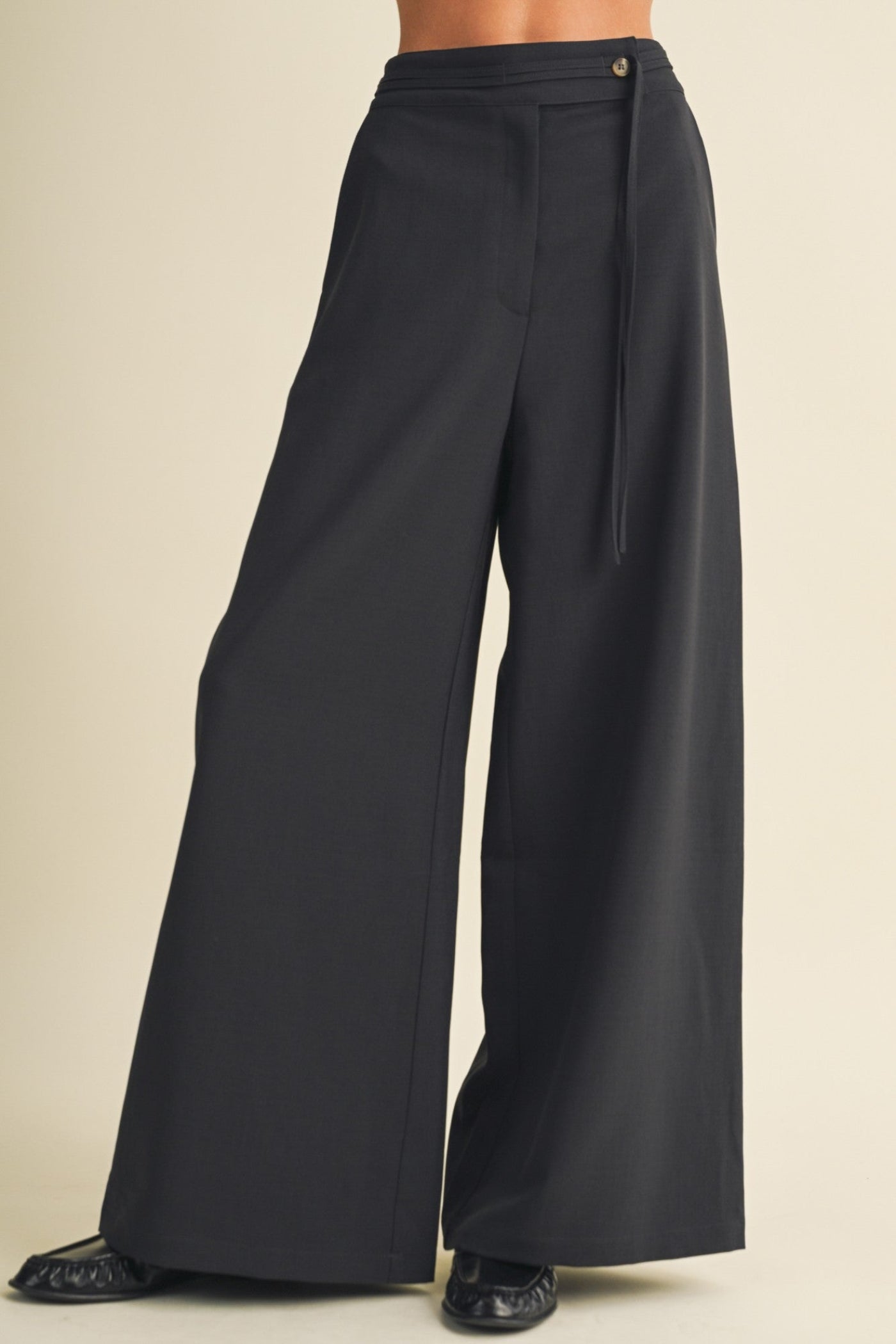 Black wide-leg trousers with a drawstring waistband, shown on a model from the waist down against a solid beige background.