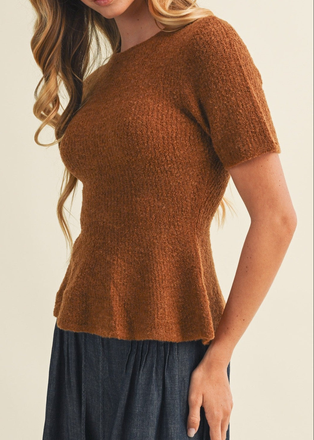 A woman wearing a short-sleeve, crew-neck knit top in a warm rust brown color, paired with a dark blue skirt, photographed against a solid light beige background.