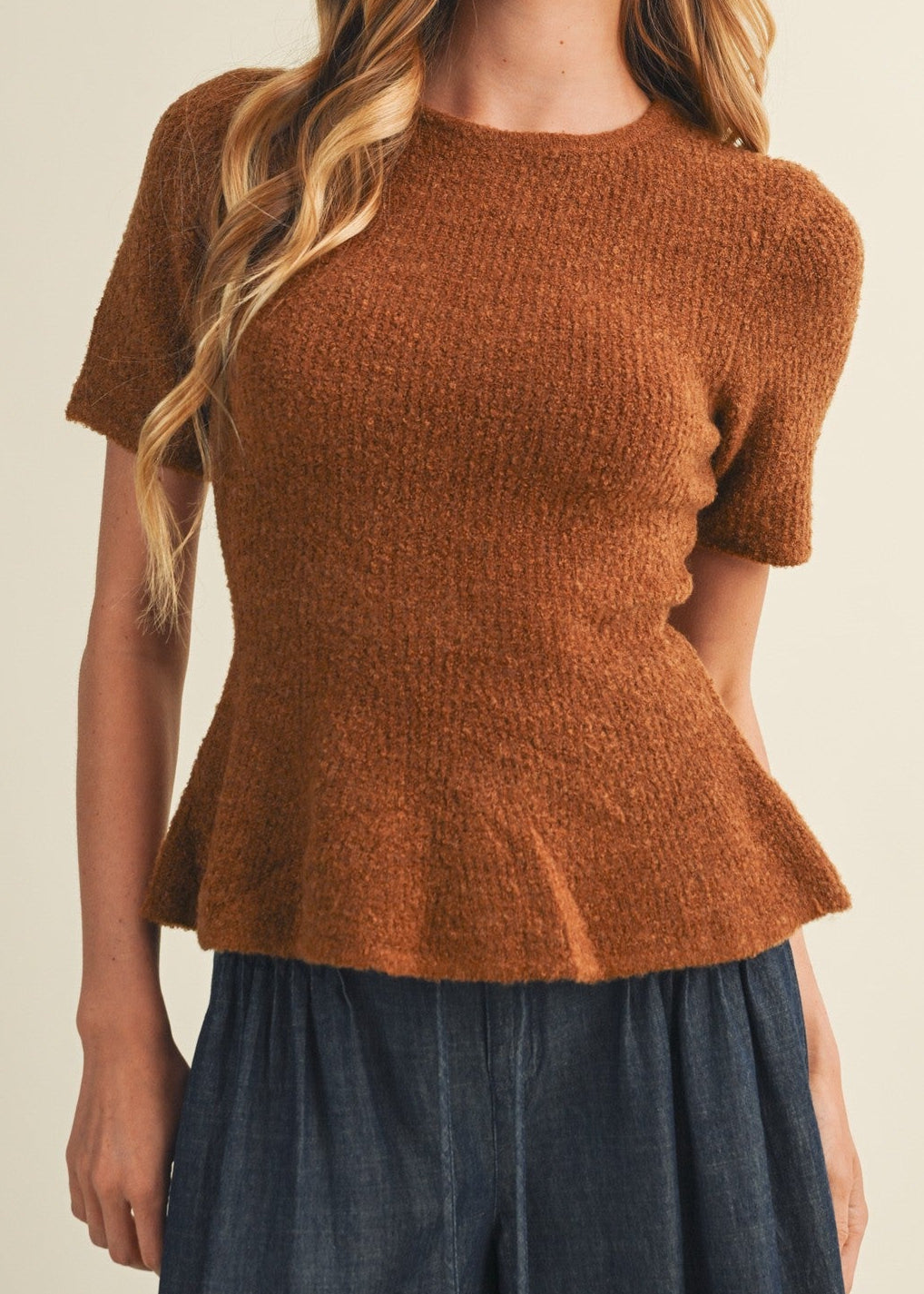 A woman modeling a short-sleeve, crew-neck knit top in a warm rust brown color, paired with dark blue wide-leg pants, against a solid light beige background.