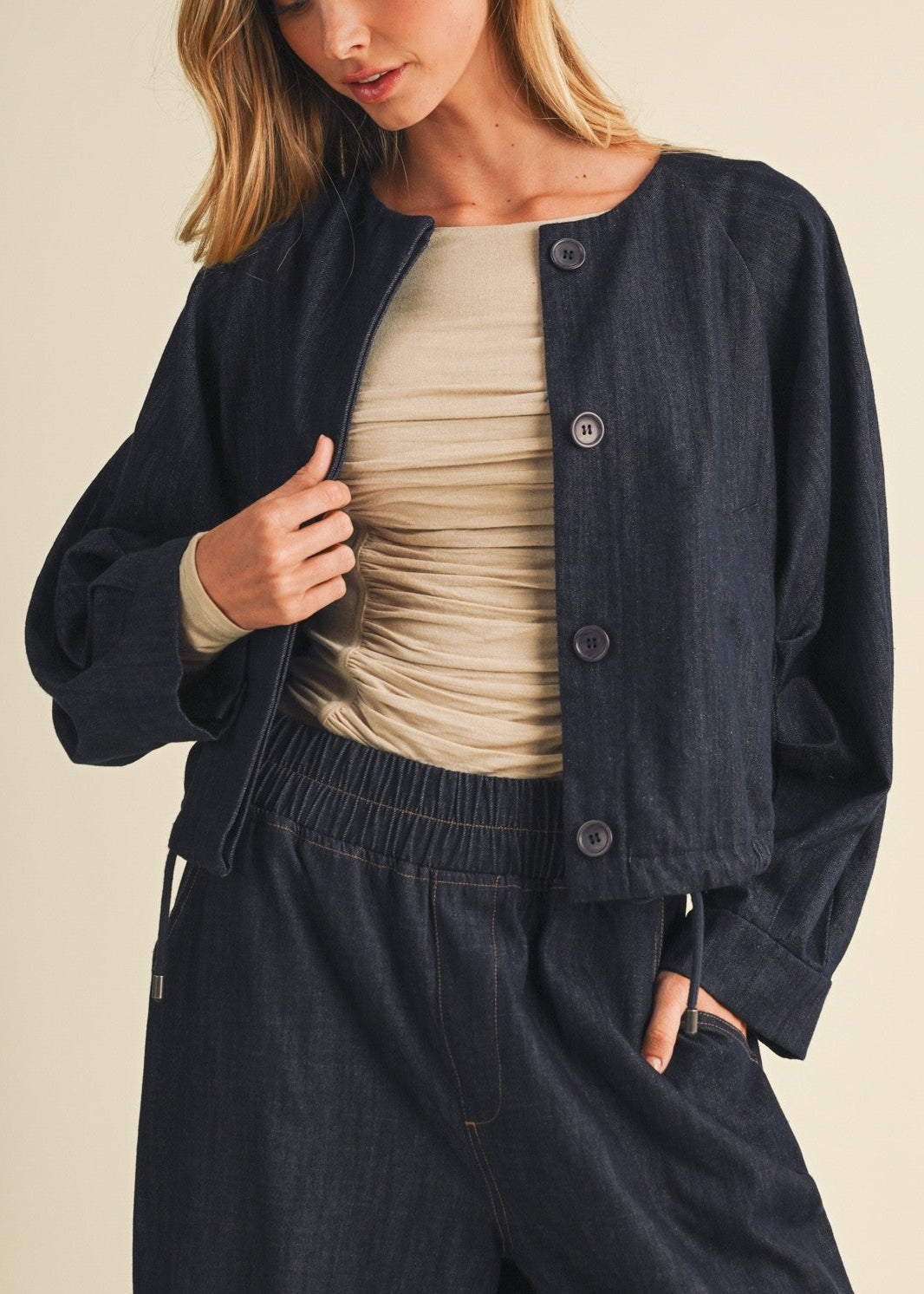 A woman wearing a dark navy blue cropped button-up jacket over a beige ruched top and matching dark navy wide-leg pants, posed against a solid light beige background.