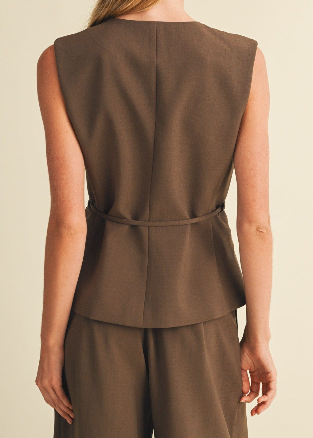 A woman shown from the back wearing a sleeveless brown peplum-style top and matching brown trousers against a plain warm beige studio background.