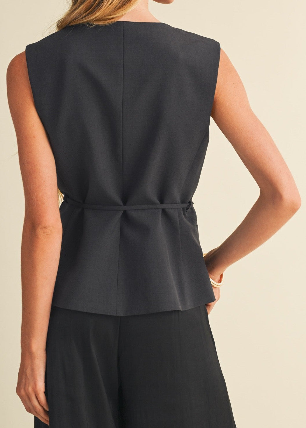 A woman shown from the back wearing a sleeveless black peplum-style top with a gathered waist seam and black wide-leg pants, against a plain warm beige studio background.